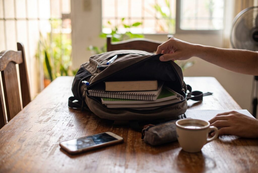 Mochila universitaria abierta con libros y cuadernos sobre una mesa de madera, junto a un teléfono y una taza de café