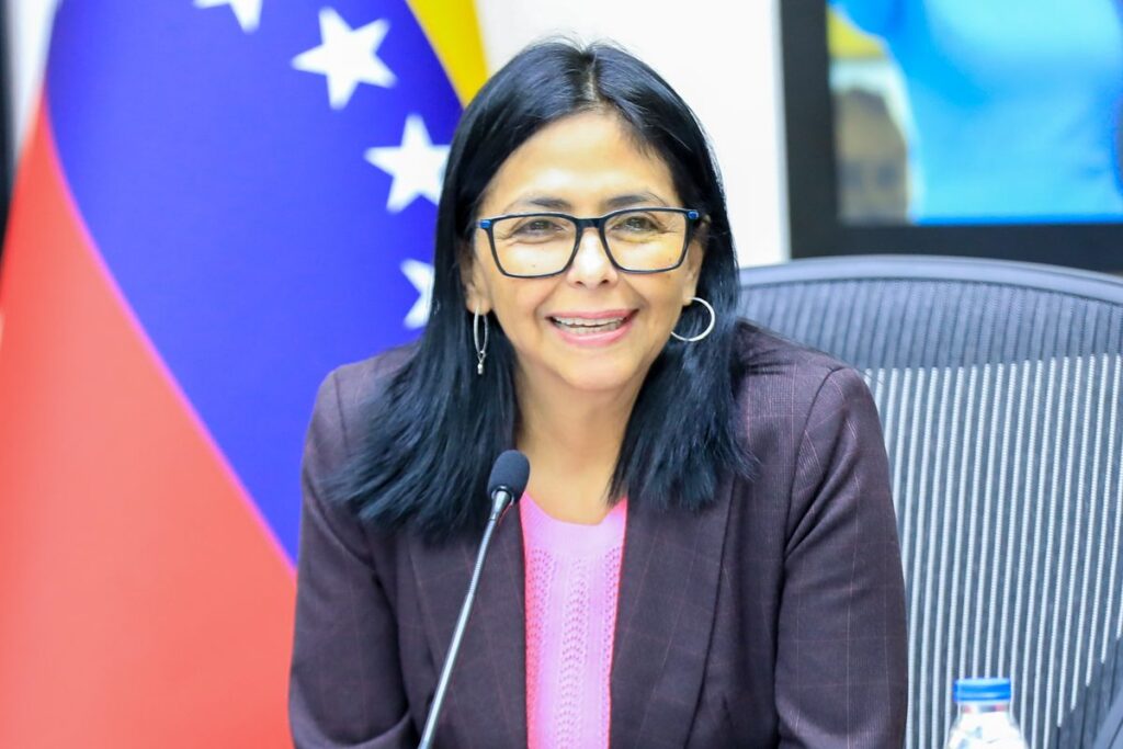 Delcy Rodríguez sentada ante un micrófono en una mesa de reuniones, con la bandera de Venezuela detrás.